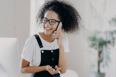 Aşırı mutlu bir kadının cep telefonuyla hoş bir şey tartıştığı, cep telefonunun yanında tuttuğu, keyifli bir ruh hali içinde olduğu, gözlük taktığı ve günlük giysiler giydiği, geniş bir odada poz verdiği, arka planda bulanıklık olan bir fotoğraf.