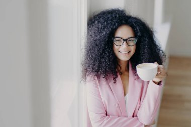 Kapalı alanda çekilen kıvırcık saçlı Afro-Amerikan kadın kahve molası veriyor, elinde beyaz bir bardak içki tutuyor, optik gözlük takıyor, resmi kıyafet giyiyor, beyaz duvarın yanında duruyor, iş dünyasında çalışıyor.