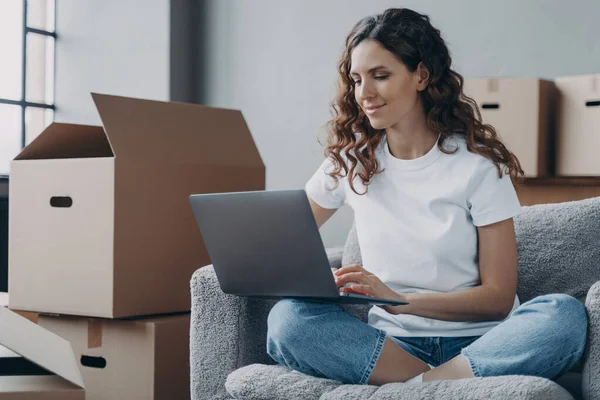 Hispanic girl sitting with laptop among cardboard boxes and smiling. Happy young woman using internet in new home. Lady moves to modern apartment. Relocation and online work concept.