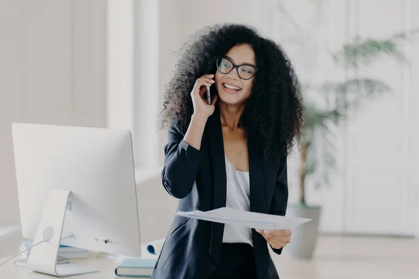 stock image Busy female manager holds mobile phone near ear, holds papers, checks report, has telephone conversation, wears glasses and elegant wear, smiles broadly, workplace with computer in background