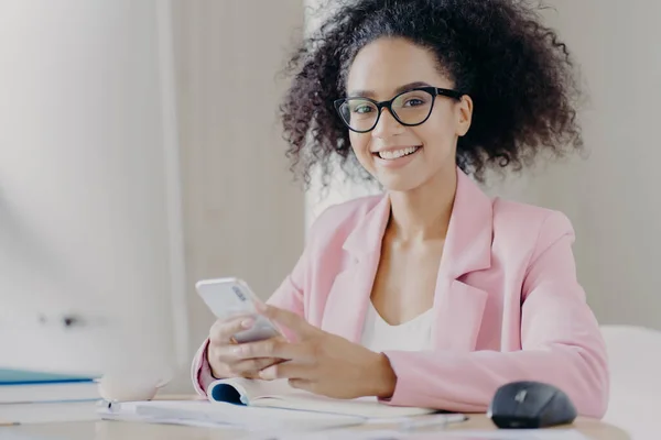 Positive curly haired woman wears spectacles, formal wear, holds modern cell phone, searches information about business news, sits at desk papers, drinks coffee, has happy expression, smiles