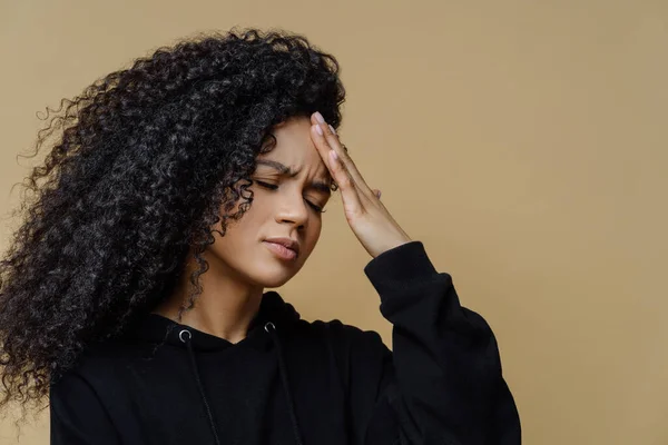 Sideways shot of displeased dark skinned woman touches forehead, suffers headache and mirgaine, squints face from pain, needs painkillers, cannot concentrate, wears black hoodie stands over beige wall