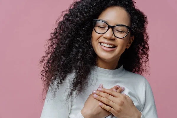 幸福诚挚的非裔美国女人紧紧抓住胸部 表达诚挚的感情 做出感恩的姿态 感激他人的帮助 咧嘴微笑 戴眼镜 — 图库照片