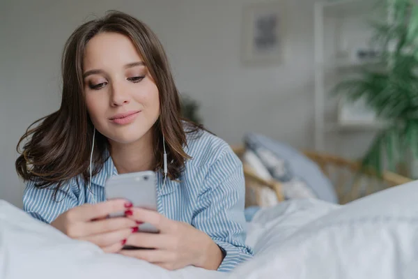 stock image Indoor shot of attractive thoughtful young woman lies in bed, uses modern cell phone, watches video or film online, enjoys good sound, dressed in pyjamas. People, bed time and relaxation concept