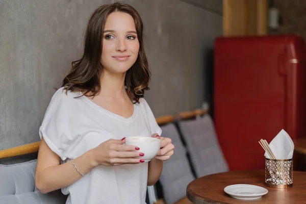 stock image Beautiful brunette woman with dreamy expression, has coffee break after work, wears white t shirt, spends free time in coffee shop, enjoys recreation time. People, lifestyle and drinking concept