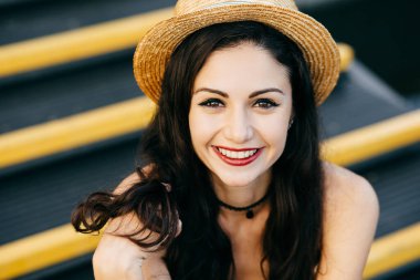 Esmer genç kadın çekici görünümü ile koyu renk gözleri, kırmızı olan dudaklar, kamera, merdiven oturan hasır şapka içine hoş gülümseyen boyalı. Güzel kadın onun olumlu duygular gösterilen