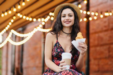 Genç kadın modeli ile koyu renk gözleri, kırmızı yazlık elbise ve eller paket servisi olan restoran kahve ve kruvasan, açık restoranda kahvaltı holding şapkası giyen dudakları boyalı. Gıda ve gevşeme kavramı