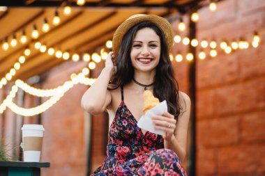 Şapka, elbise ve kolye, lezzetli kruvasan neşeli göz olan holding giyen görünüm, çekici güzel kadın siyah saçlı. Mutlu genç güzel kadın terasında Kahvaltı