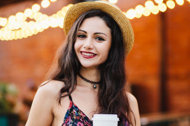 Holding kağıt bardak kahve, hasır şapka, kolye ve elbise giyen genç bir bayanla makyaj, hoş restoranda poz kameraya gülümseyen. Memnun ifade café'de oturan kadınla