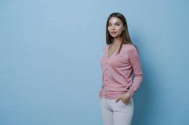 Güzel, koyu renk saçlı genç Avrupalı bir kadının yan fotoğrafları şık bir kazak giyer, beyaz pantolonun cebinde el ele tutuşur, mavi arka planda poz verir, reklam içeriğiniz için boş bir alan yaratır..