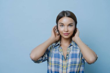 Güzel, koyu renk saçlı Avrupalı bir kadının portresi sağlıklı, parlak bir cilde sahip, makyajlı, kareli gömlek giymiş, doğrudan kameraya bakıyor, mavi arka plana karşı duruyor, iyi bir ruh hali içinde.