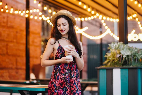 stock image Dreamy woman with dark hair wearing hat and dress with flower print looking down while having some thoughts in her mind, holding paper cup with fresh cappucino standing at terrace of cozy cafe