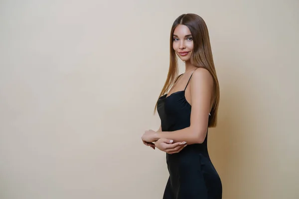 stock image Beautiful slim woman stands in profile, wears elegant black dress, has long straight hair, looks with charming expression at camera, poses against beige background, copy space for your information