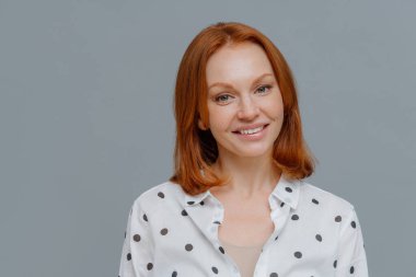 Zarif kızıl saçlı kadın kameraya nazik, dişlek bir gülümsemeyle bakıyor, sağlıklı çilli bir teni var, iyi haberler duymaktan mutlu, şık bir gömlek giyiyor, gri duvarın üzerinden izole edilmiş, metnin için kopyalama alanı var.