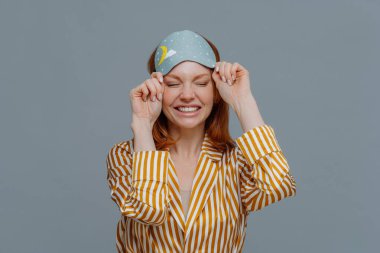 Günaydın, yaşam tarzı. Gülümseyen çilli kadın göz bağını çıkarır, sarı çizgili pijama giyer, hoş rüyalar gördükten sonra gülümseyerek uyanır, rahatlamış ve tazelenmiş hisseder, gri duvarın üzerinde poz verir.