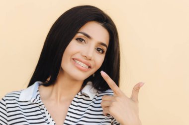 Portresi yakından esmer kadın, koyu renk gözlü, güzel yapmak kadar memnun, ağzında gösterir, geniş bir gülümseme var, beyaz dişler diş telleri ile gösterir, boş duvara izole güzelliği gösterir