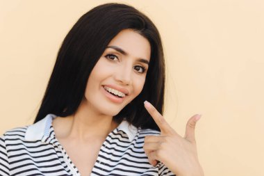 Kadın lüks siyah saçlı, beyaz dişli destekleri ile yakışıklı, ağız ile ön plana gösterdiğine parmak, diş teli, diş hekimi, stüdyo arka plan üzerinde izole ziyaretinden sonra memnun tanıtır