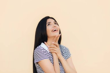 Neşeli muhteşem genç kadın modeli mutlu yukarı bakar ve tutar el altında çene, fark bir şey keyifli, rahat kıyafetler giymiş giyer ışık arka plan üzerinde izole dişler, diş teli.