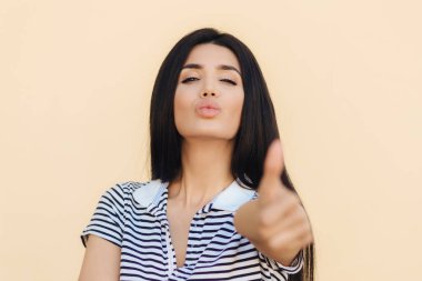 Tamam o zaman! Koyu saçlı güzel kadın güvenle kameraya benziyor, başparmak jest gibi gösterildiği gibi yükseltir, bej renkli arka plan üzerinde izole gündelik kıyafeti giymiş bir şey onayladı.