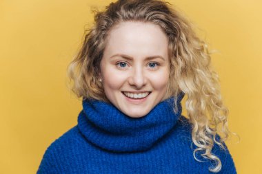 Hafif gevrek saçlı mutlu mutlu kadın sıcak mavi Kazak giyer, geniş bir gülümseme ve beyaz dişler sarı arka plan üzerinde izole başarılı gün sonra memnun vardır. Olumlu duygular