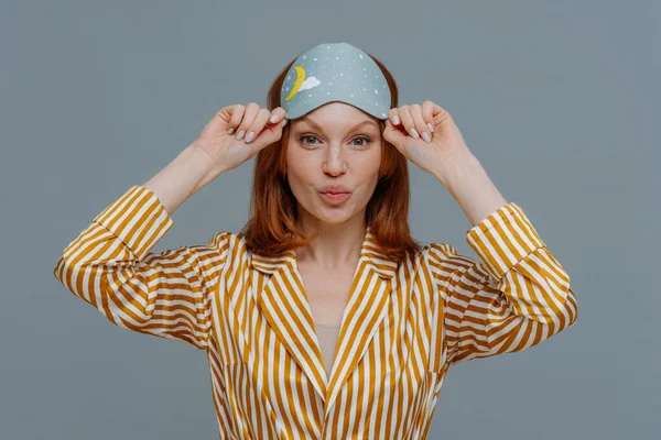Beautiful freckled ginger woman wears sleepmask and striped pajama, keeps lips folded, has healthy skin, going to have sleep at home, enjoys day off and lazy day, isolated over grey background