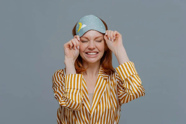 Good morning, lifestyle. Smiling freckled woman takes off blindfold, wears yellow striped pajama, awakes with smile after seeing pleasant dreams, feels relaxed and refreshed, poses over grey wall
