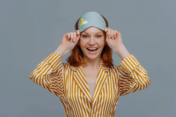 Ginger female model wears sleepmask and striped pajamas, awakes in good mood, realizes today is weekend, can stay in bed for long time, smiles positively, isolated on grey background. Sleeping