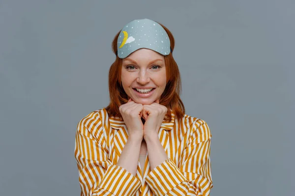 Headshot of attractive woman keeps hands under chin, has ginger hair, pleasant toothy smile, wears yellow nightwear and sleepmask, looks directly at camera, isolated over grey background. Wake up