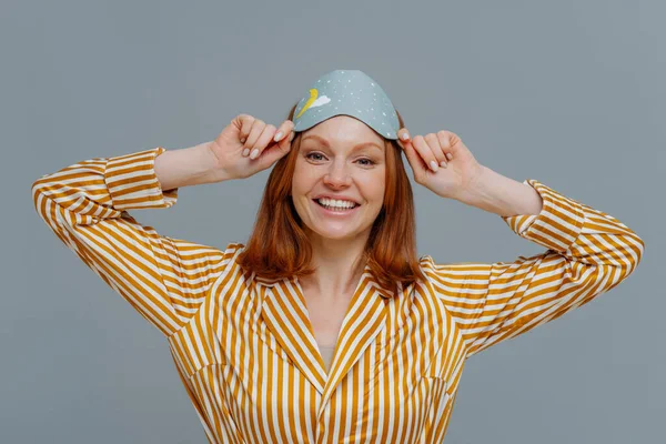 Positive redhaired young female wears eye mask for sleep on head, smiles gladfully, dressed in home wear, has sweet dreams at night, isolated over grey background, enjoys peaceful atmosphere