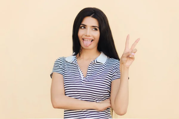 Positive funny female makes peace sign with hand, shows tongue, being in good mood, has long dark hair, has healthy skin, wears casual clothes, isolated over studio background. Body language
