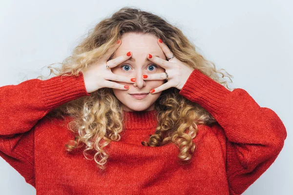 Horizontal portrait of mysterious blue eyed female with curly light hair, covers face with hands, dressed in loose red sweater, isolated over white background. Surprisment and people concept