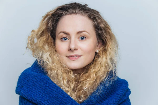 Portrait of pleasant looking curly woman with pleased delighted expression, has curly hair, wears casual warm sweater, looks directly into camera, isolated over light blue studio background.