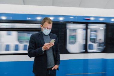 Metro istasyonunda pozlar veren, toplu taşıma araçlarıyla seyahat eden, rotayı kontrol etmek için modern cep telefonu kullanan, koronavirüs veya gribe karşı tıbbi koruyucu maske takan erkek işçilerin yatay görüntüsü. Sağlık tehlikesi