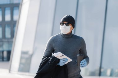 Balıkçı yaka ve şapka giymiş, salgın sırasında koruyucu maske ve lastik eldiven takan, kahve içen ve elinde gazete tutan beyaz adamın yatay görüntüsü.