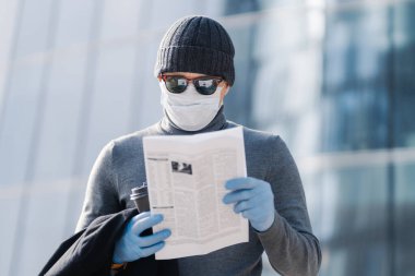 Genç adam Coronavirus belirtileri hakkında makale okuyor, koruyucu maske ve lastik eldiven takıyor, sokakta kahve ile poz veriyor, kurallara uyuyor veya karantinaya alınıyor. Covid-19 koruma kavramı