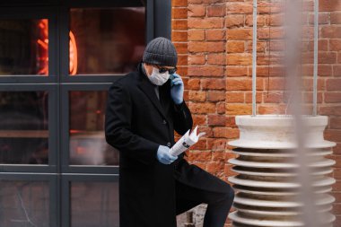 Erkek girişimci, akıllı telefondan çalışan, koruyucu tıbbi maske takan, eldiven takan, kağıt tutan, tuğla duvar pozu veren kişilerden bahsediyor. Salgın virüsü. Roman Coronavirus konsepti. Güvende ve sağlıklı kalın.