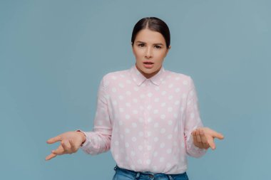 Makyaj ile Pretty koyu saçlı kadın, elleri yayılır ve şüpheyle görünüyor, yanlış anlama hissediyor, polka nokta gömlek ve kot giyer, karar veremez, mavi stüdyo arka plan üzerinde kapalı pozlar