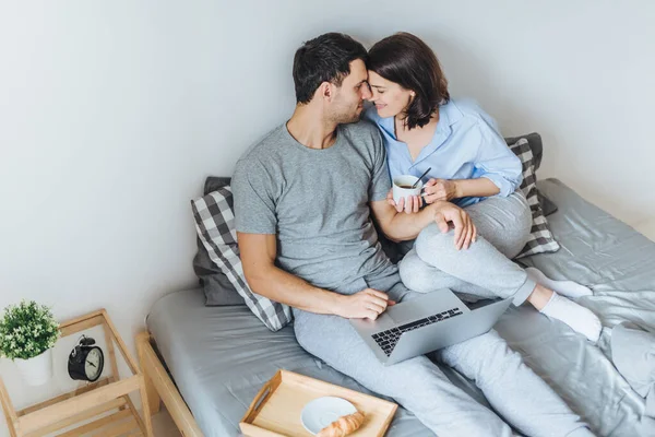 stock image Lovely romantic couple going to kiss, look at each other as sit on bed, use laptop computer for watching films, drink coffee, enjoy togetherness and relaxation in bedroom. Relationship concept