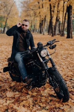 Düşünceli motorcunun dikey görüntüsü güneş gözlüğü takıyor, parkta motosiklet sürüyor, sokakta poz veriyor, şık kıyafetler giyiyor, boş zamanlardan ve hobilerden hoşlanıyor, doğaya hayran, güneşli sonbahar havasından hoşlanıyor.