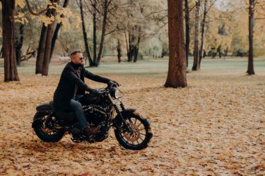 Kendine güvenen erkek motorcu motosiklet sürüyor, tek başına seyahat etmekten hoşlanıyor, koruyucu güneş gözlüğü takıyor, rahat kıyafetler giyiyor, sonbahar parkında ağaçların arasından geçiyor. Sonbahar sezonu, hobi, yaşam tarzı.