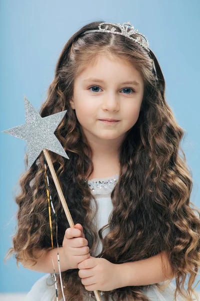 Small kid angel has long crisp hair, blue eyes, holds magic wand in form of star, poses over blue background, has appealing appearance. Little princess waits for miracle, believes in fairy tale