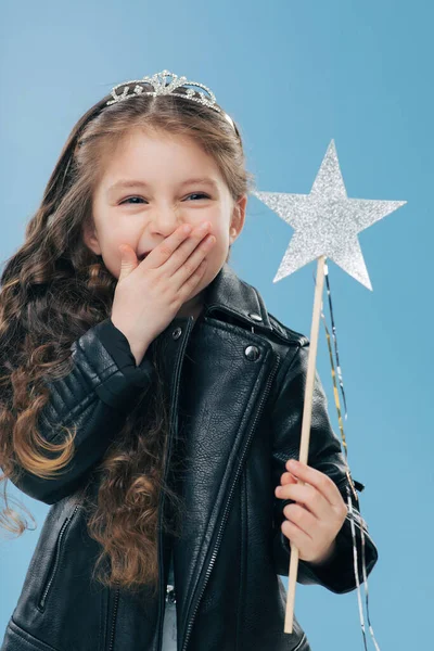 Overemotive positive small child covers mouth with one palm, wears fashionable black leather jacket and crown, holds magic wand in hand, isolated over blue background. Female kid poses indoor