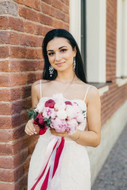 Güzel esmer kadın, çekici bir görünüm, küpe, beyaz gelinlik giyer ve güzel buket tutar, duvara karşı duruyor. İyi görünümlü gelin özel durum için mutlu