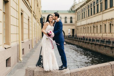 Sevgi evli çift öpücük tutkuyla Köprüsü'nde antik şehirde Nehri yakınında dururken kutlamak creat mutlu olmak onların düğün onların dostu aile. Romantik yeni evliler açık havada poz