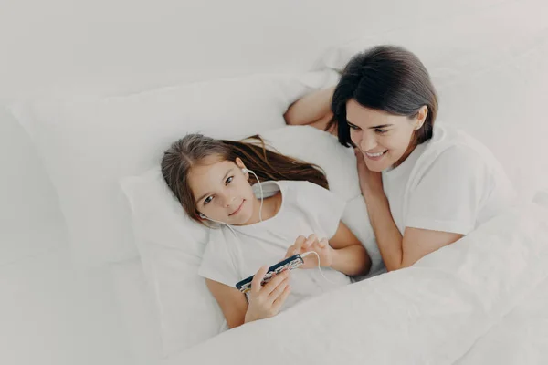 Caring mom looks attentively at her little daughter who wakes up early, listens music in earphones, searches new song in playlist of modern smartphone, lie under soft white blanket. Top view