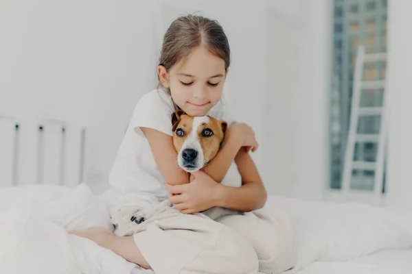 Indoor shot of happy small child embraces favourite pet, dressed in pyjamas, expresses love to her dog, plays with animal after awakening, poses on soft bed. Children and domestic animals concept