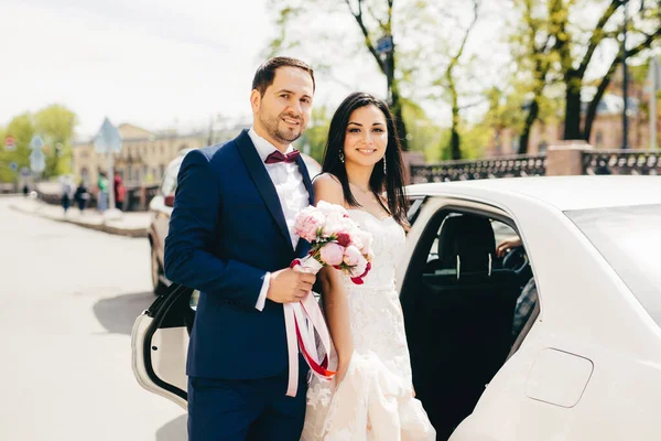 Lovely married couple stand next to each other near car, going in restaurant to celebrate their wedding, have party, invite friends and relatives. Newlyweds pose outdoor after registration of marriage