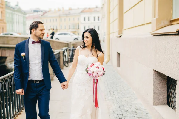 stock image Attractive female with dark hair, wears white dress, holds bouquet, keeps bridegroom`s hand, look at each other with great love, have walk across ancient city on bridge. Positive emotions concept