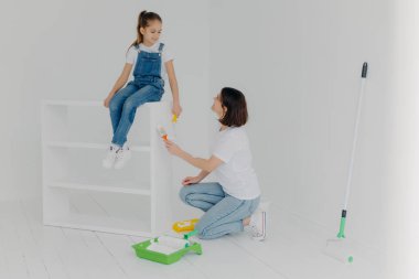 Memnun esmer anne çocuğa bakıyor, modern dairede mobilya boyuyor, beyaz duvarlı boş bir odada poz veriyor, evi yenilemekle meşgul. Aile, onarım ve resim konsepti