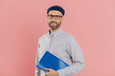 İnsanlar ve eğitim kavramı. Sakallı Genç beyaz adam öğrenci ders kitabı tutan, son sınav için hazırlar, Gözlük, şapka ve resmi gömlek, pembe bir arka plan karşı yan duruyor giyer